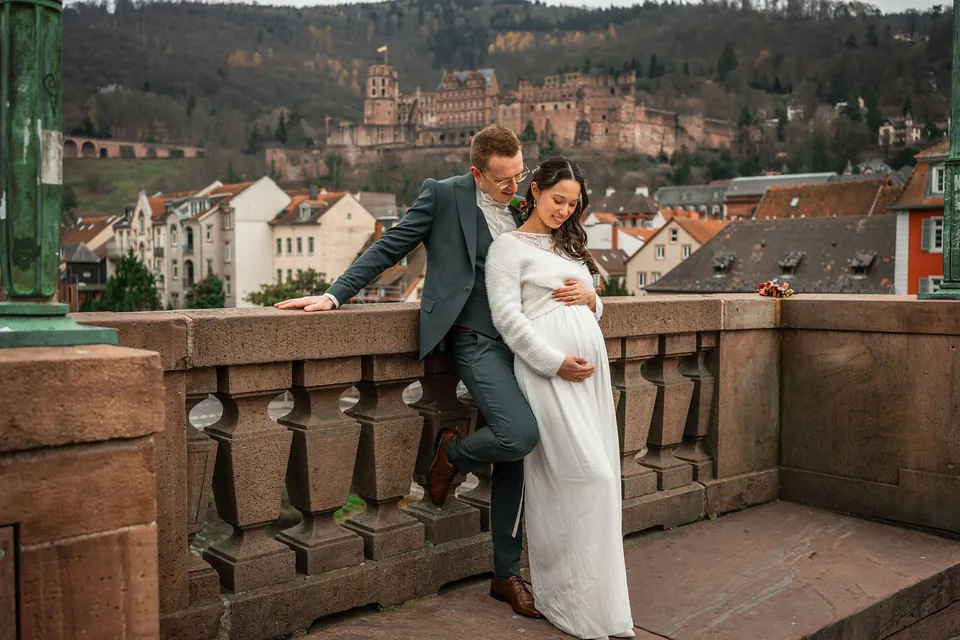 Jens lehnt an der Sandsteinbrüstung der Alten Brücke, Kim im Arm, darüber das Heidelberger Schloss, Brautpaarshooting in Heidelberg