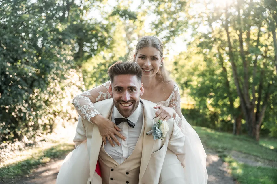 Robin trägt Melanie lachend huckepack, hinter ihnen die Sonne zwischen den Bäumen, Hochzeit in der Schleifenmühle Dettenheim-Rußheim bei Karlsruhe