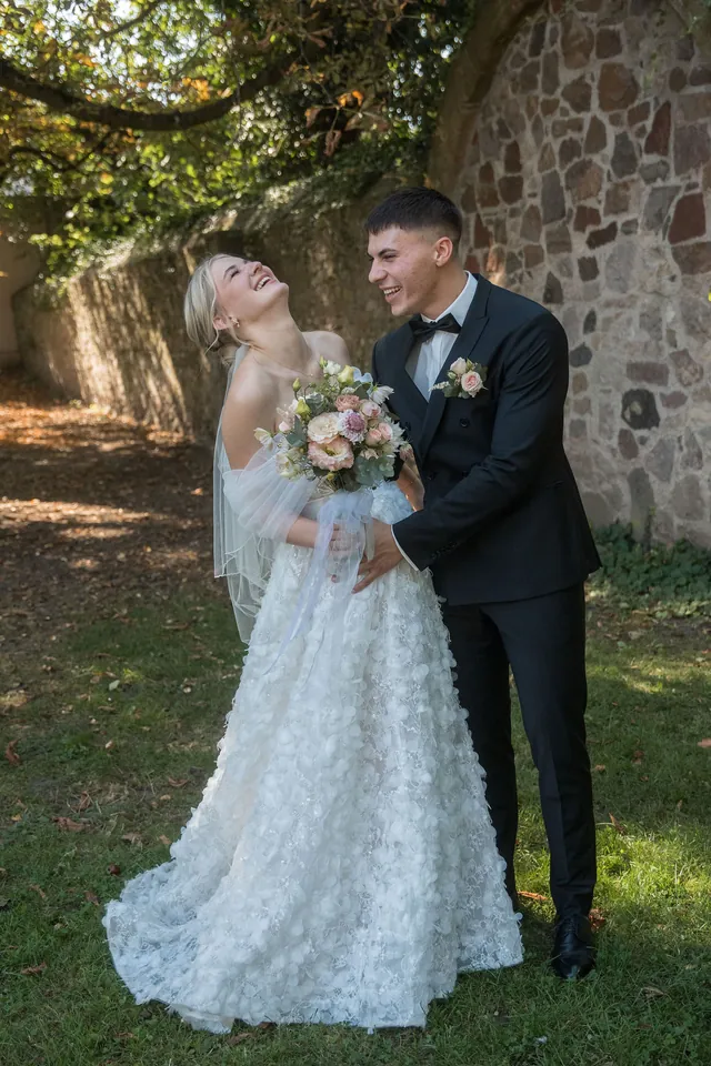 Braut lacht im Arm des Bräutigams an der Steinmauer, Hochzeit am Schloss Schwetzingen